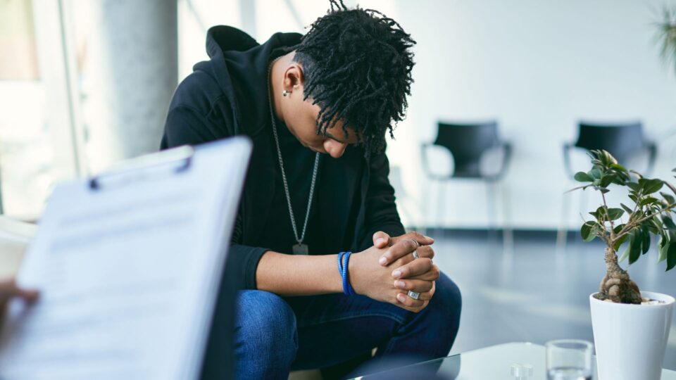 Young African American man wearing military dog tags over black sweatshirt sits with bowed head in therapist office. Clipboard showing at edge of photo.