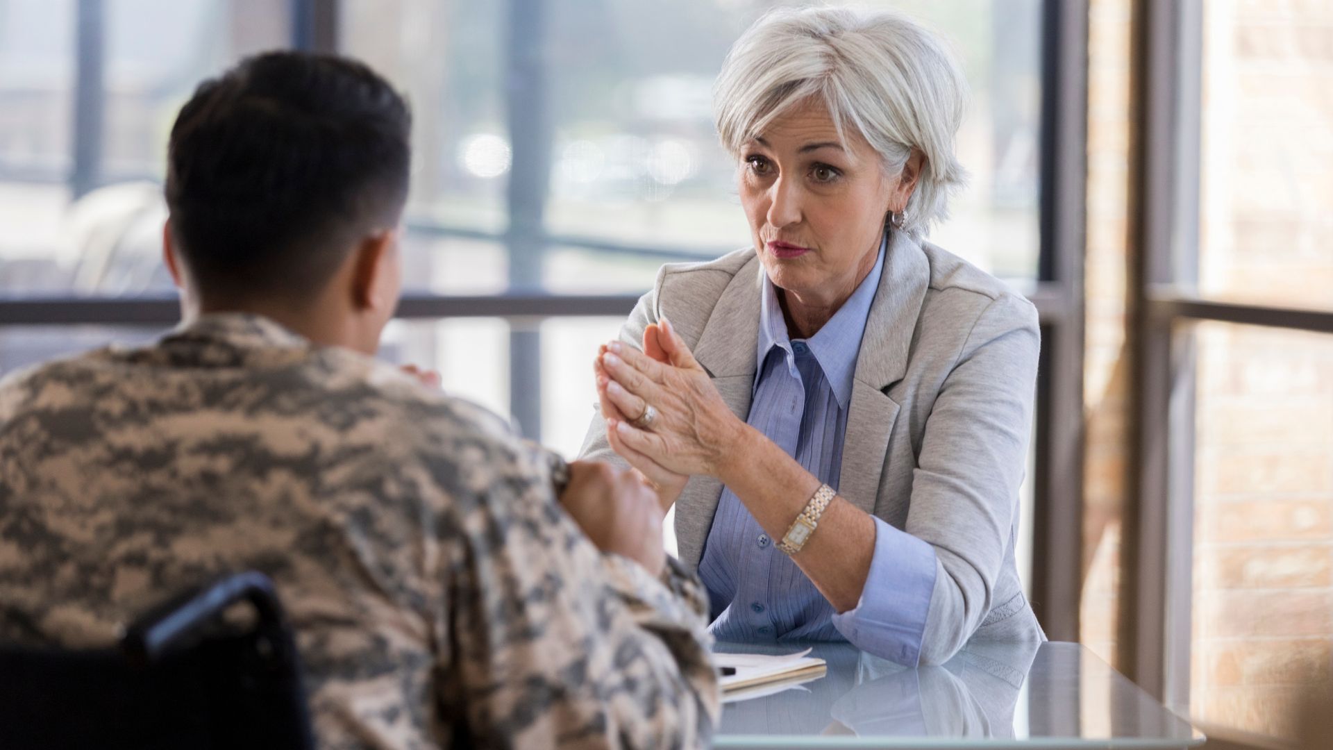 Older woman therapist with silver hair talks with military man sitting in a wheelchair.